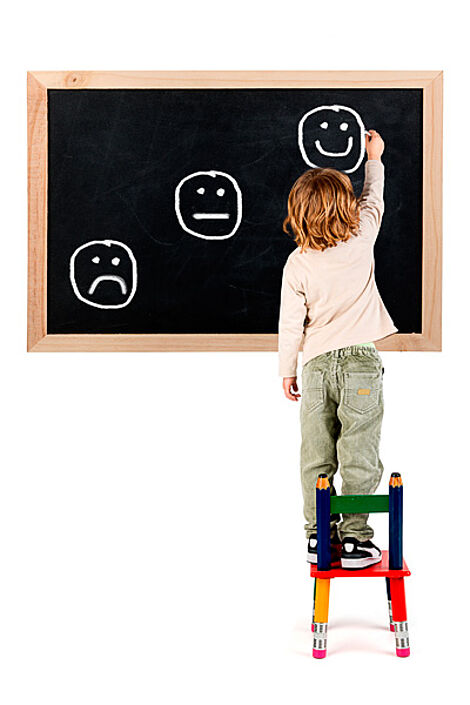 Kind auf Stuhl malt immer stärker lächelnde Smileys auf Tafel