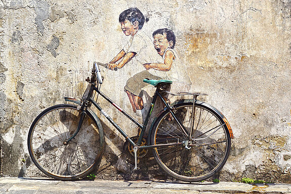 Fahrrad steht vor einer Wand, auf die Wand sind ein Mädchen und ein Junge gemalt, die auf dem Fahrrad fahren.