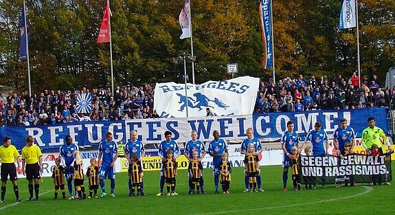 Fan-Initiative Oldenburg mit Bannern "Refugees welcome" im Stadion, davor Fußballmannschaft